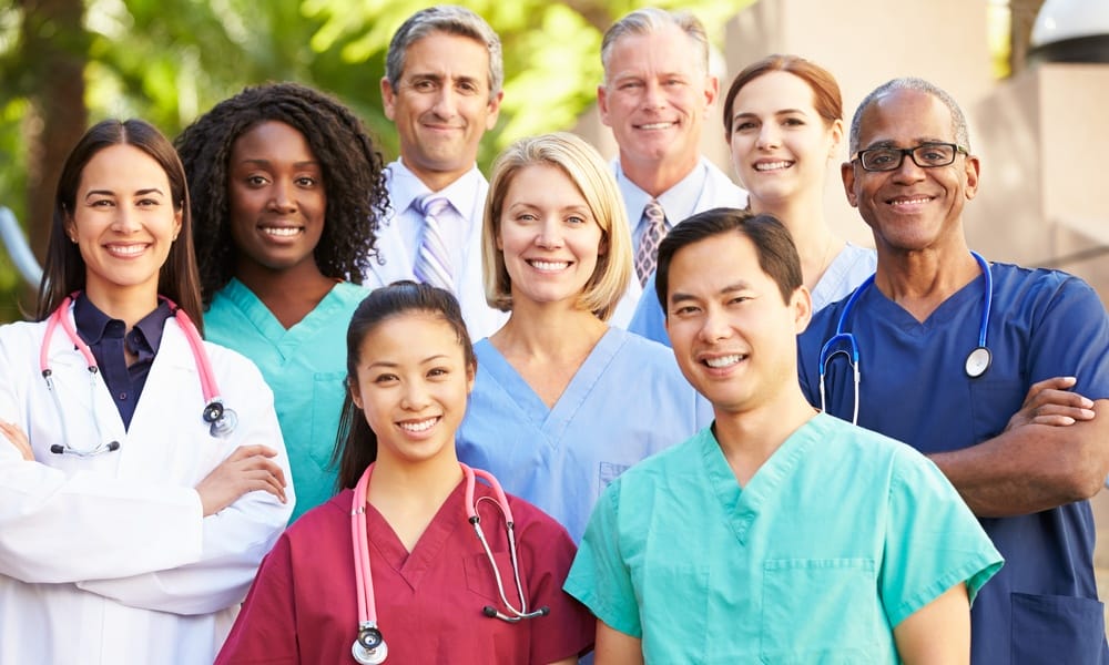 A group of smiling people dressed in different colored scrubs. Some have stethoscopes around their necks.