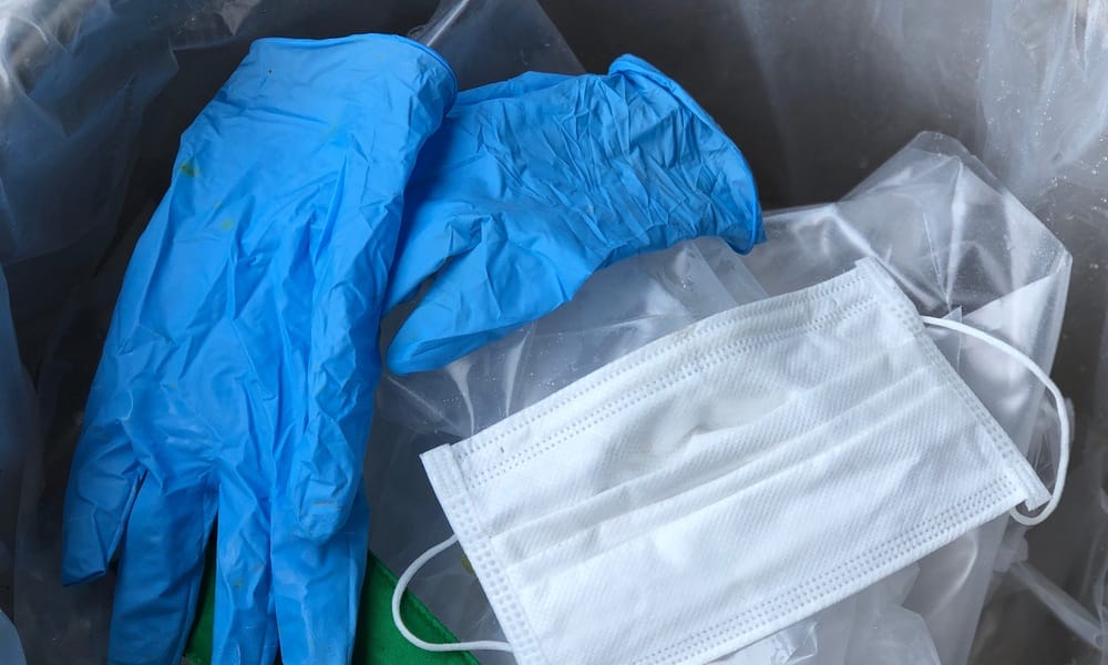 A hospital waste bin with a can liner and waste inside the bin. There's a pair of blue latex gloves and a used face mask.