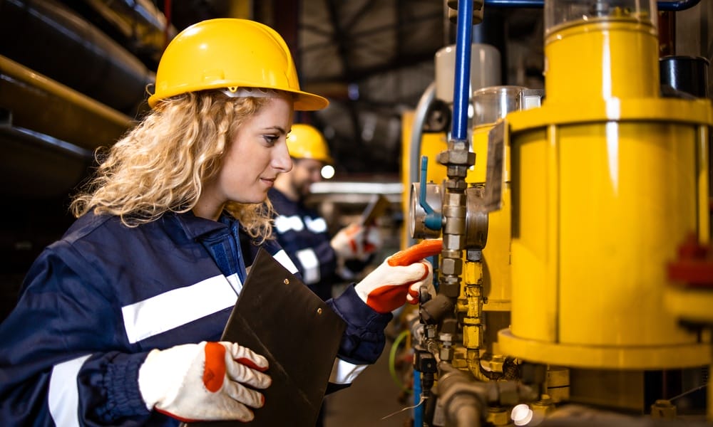 A worker wearing gloves and a hard hat is examining an industrial pipeline in a factory while holding a clipboard.
