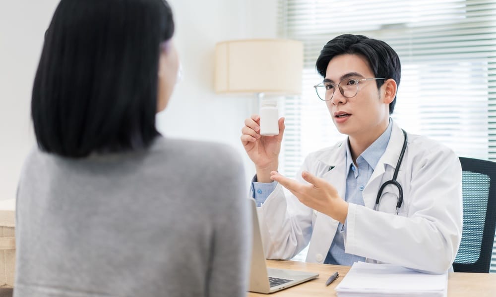 A male doctor wearing a stethoscope holds up a white pill bottle while talking to a woman sitting across him.