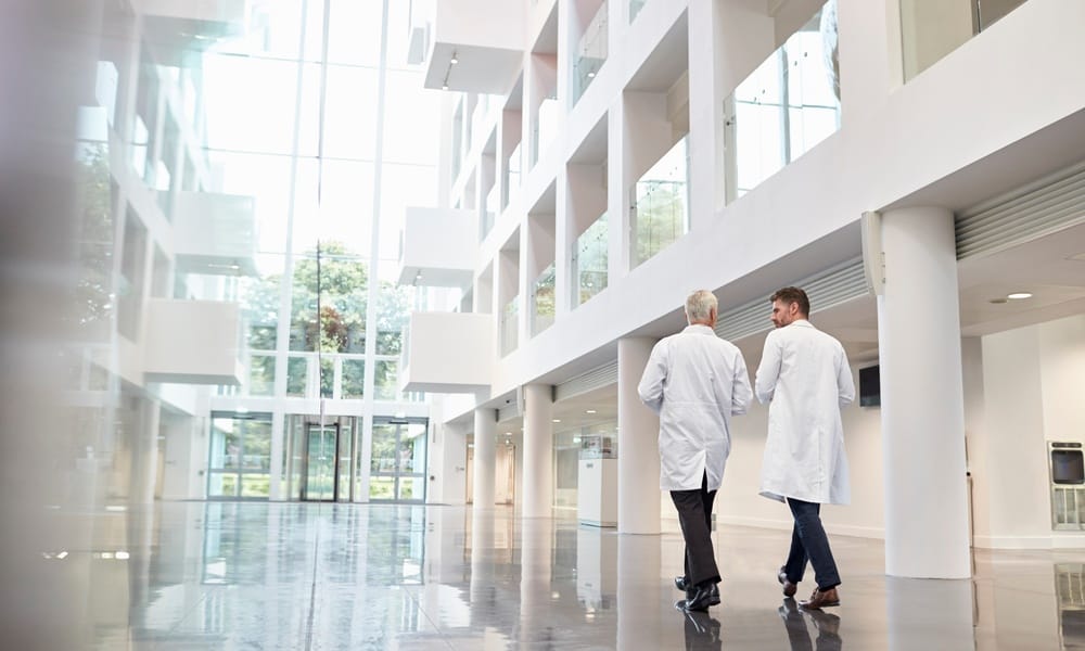 Two men in lab coats walking and talking inside a multistory building with white walls and a tiled floor.