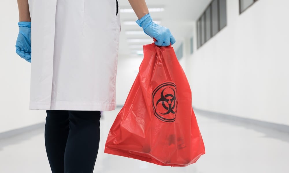 A doctor wearing blue gloves and a white coat walks down a hospital hallway, holding a red biohazard bag.
