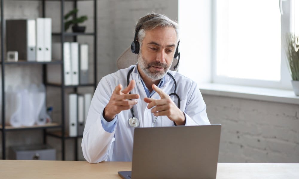 A doctor wearing a headset and white lab coat with a stethoscope around his neck sits at a desk with a laptop.