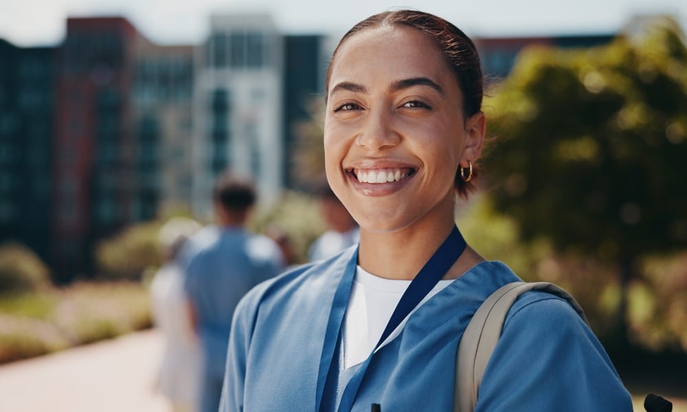 A Black woman healthcare professional smiling for the photo in her scrubs. She's standing outside the hospital.