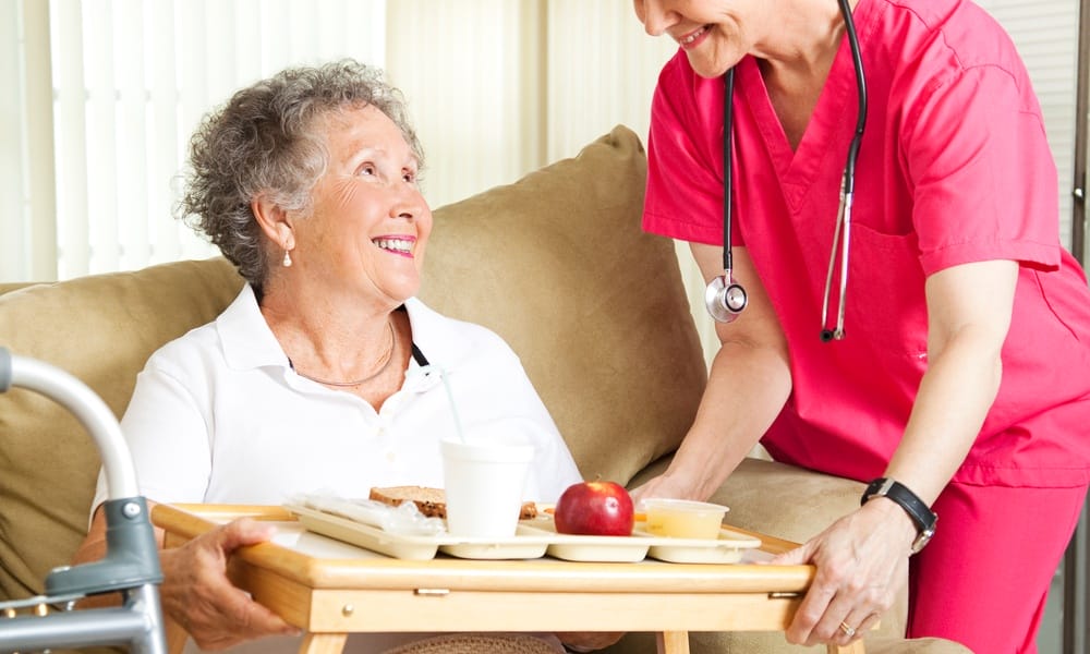A senior woman sitting on the couch with a TV tray in front of her and a hospice nurse placing lunch on the tray.