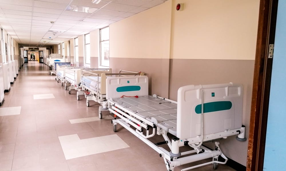 A long hospital corridor lined with movable bed frames. Several windows run along the wall, letting in natural light.