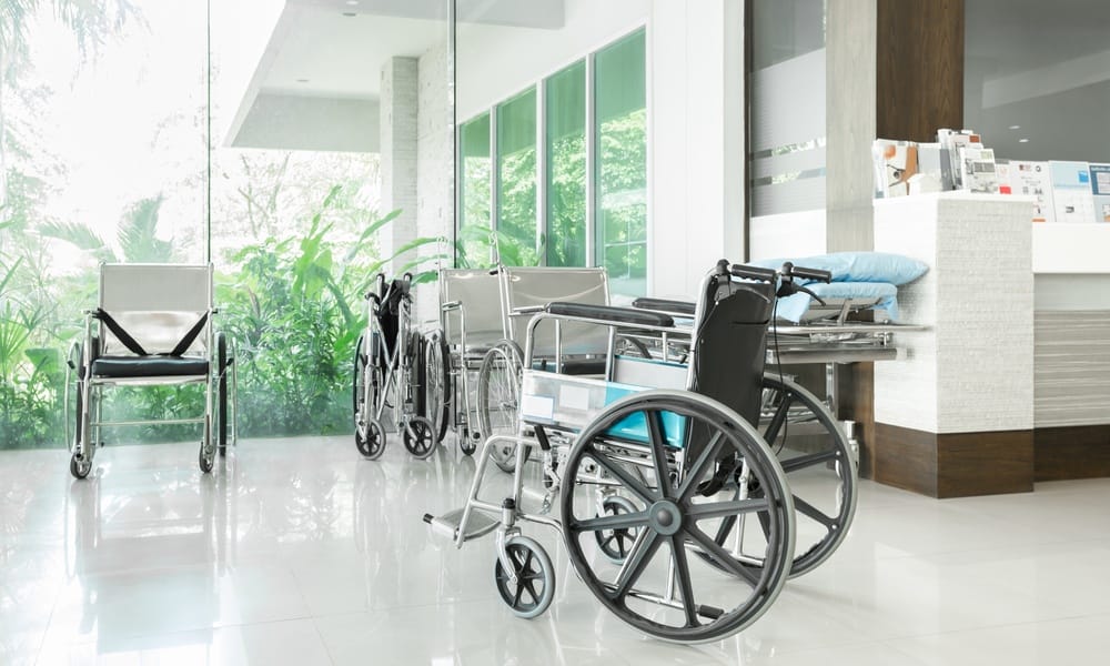 Several empty wheelchairs sit in a hospital hallway in front of a window wall near a counter with various brochures.