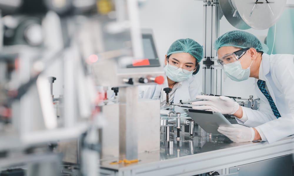 Two people wearing hair nets and gloves observe various lab equipment. One of them is holding a tablet.