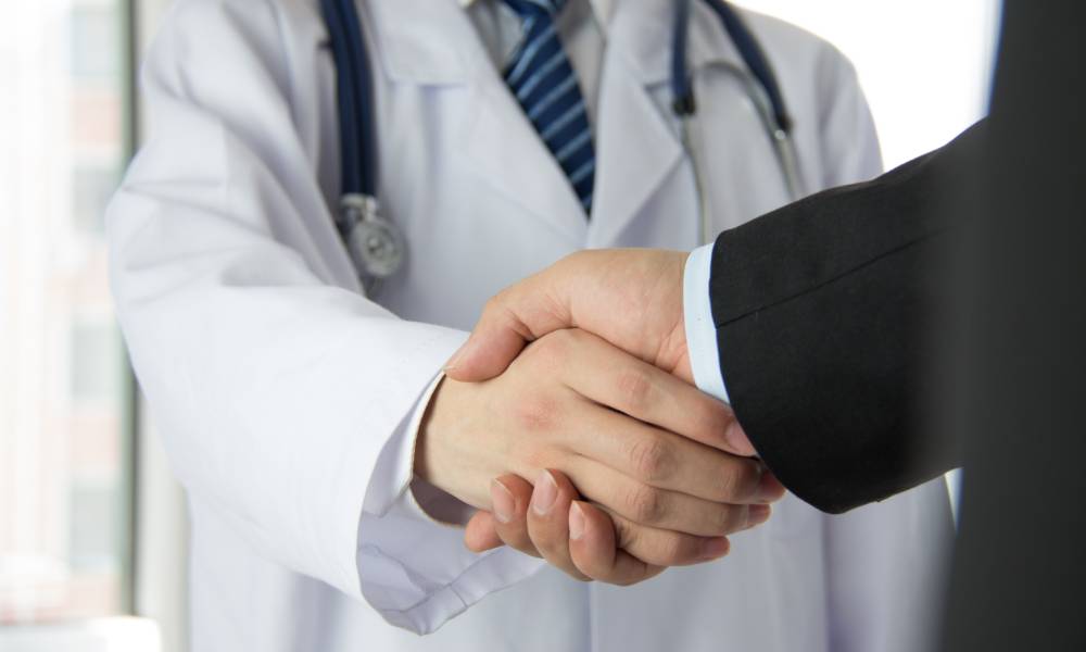 A person wearing a white medical coat and a stethoscope shaking hands with a person wearing a black business suit.