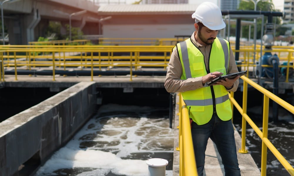 A service worker in a vest and hard hat is using a tablet. He's standing on a catwalk at a water treatment plant.