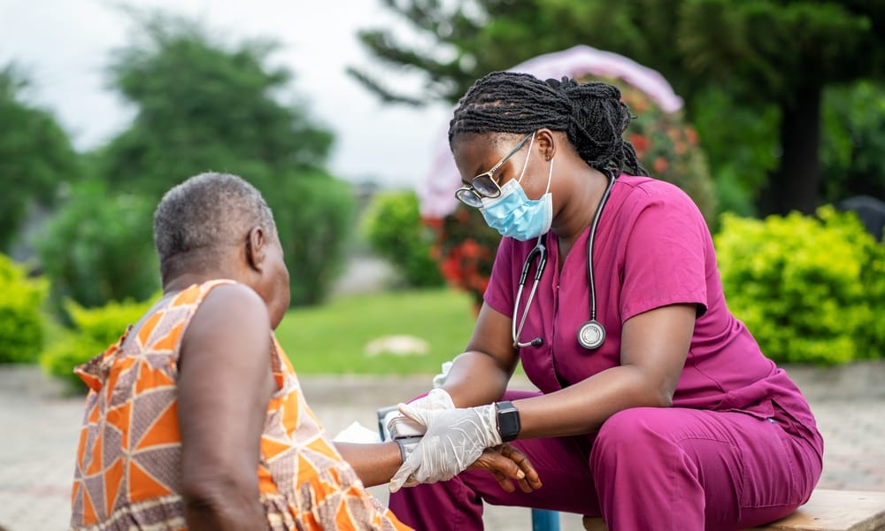 A female healthcare worker in purple scrubs and a face mask examining an elderly patient's arm outdoors.