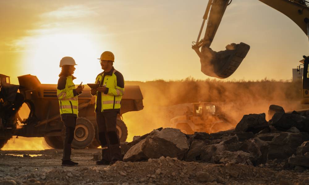 Two construction workers standing in the middle of a site. There is an excavator moving rubble in the background.