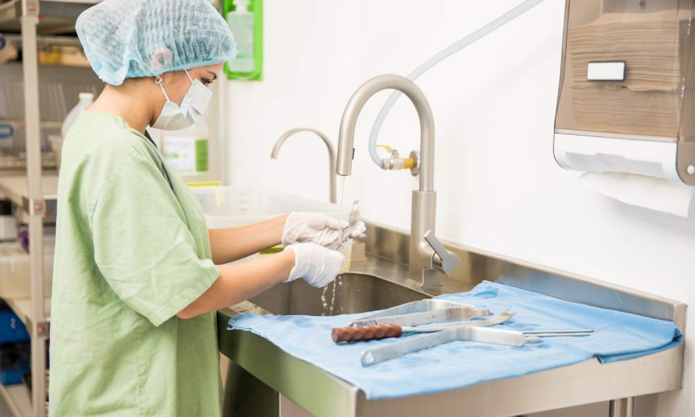 A medical professional woman in green scrubs and a hair net is sterilizing medical instruments in a hospital.