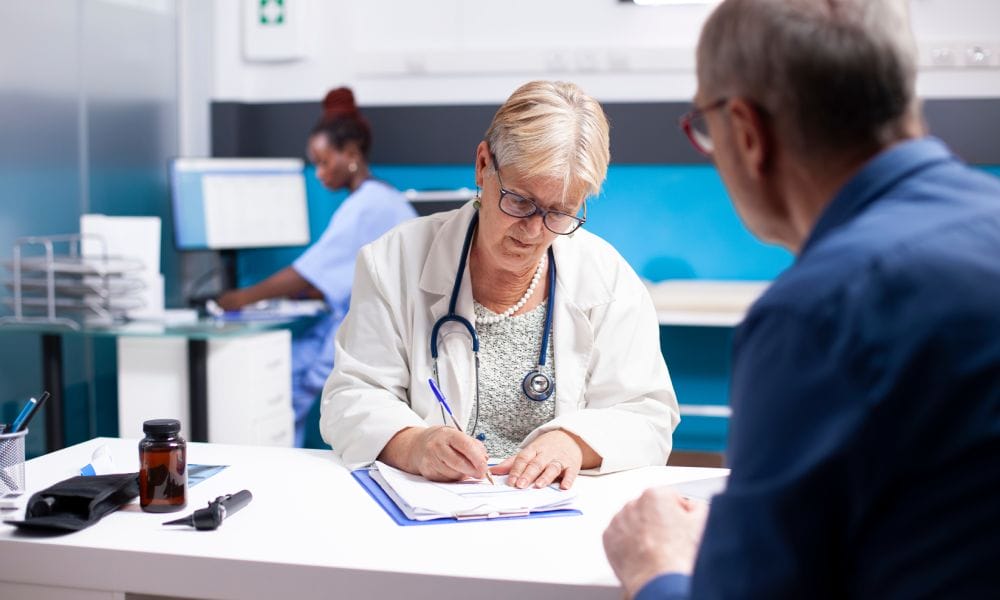 A mature woman doctor, wearing red glasses, listens to an older man's symptoms and notes them on a clipboard.