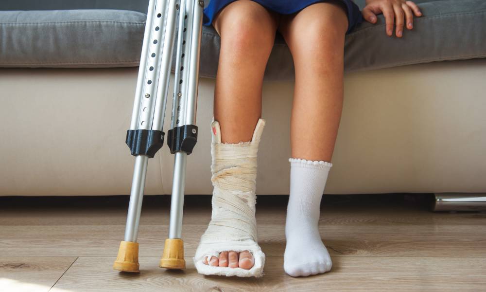 A child sitting on a couch while holding silver crutches at their side. One of their feet is current in a protective cast.
