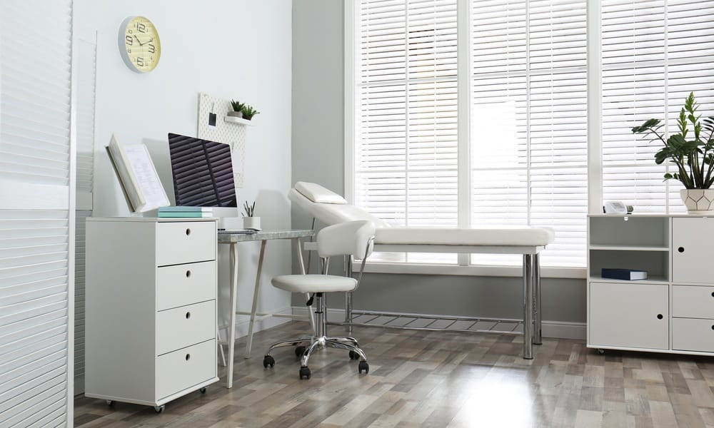 A modern exam room featuring a neutral color palette, with gray and white walls, wood flooring, and sleek white furniture.