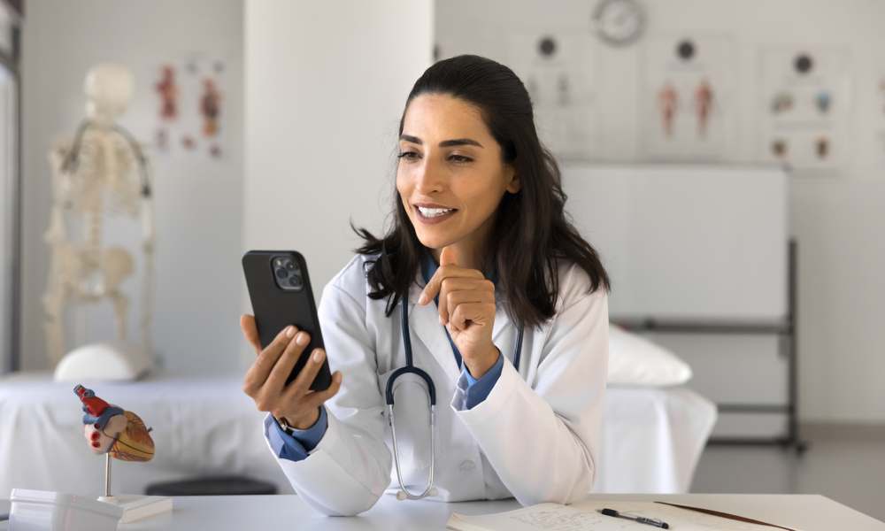 A doctor, wearing a stethoscope and medical attire, sits at her desk and smiles as she looks down at her cell phone.