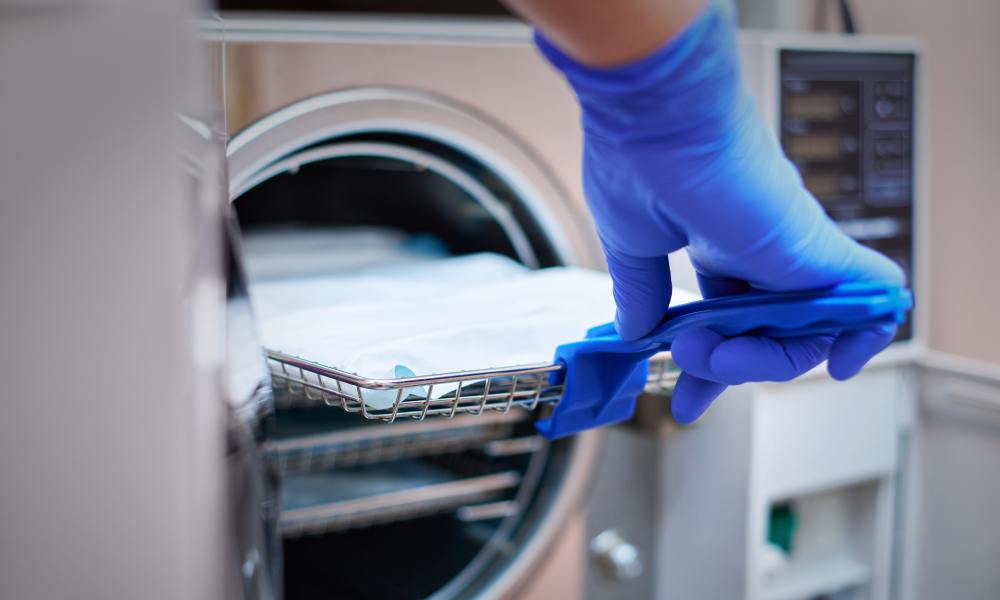 A medical professional pulling out a metal tray from an autoclave with a plastic tool. They are wearing blue latex gloves.