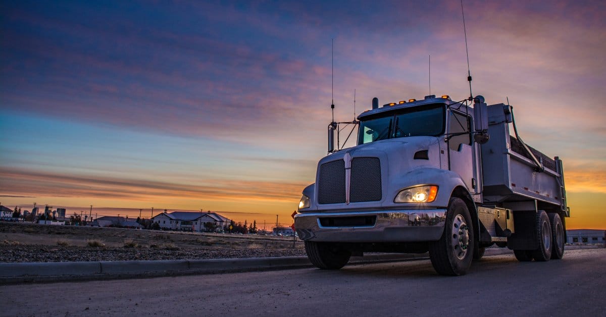 A semi-truck driving through a rural highway at sunrise. There are residential homes in the distance.