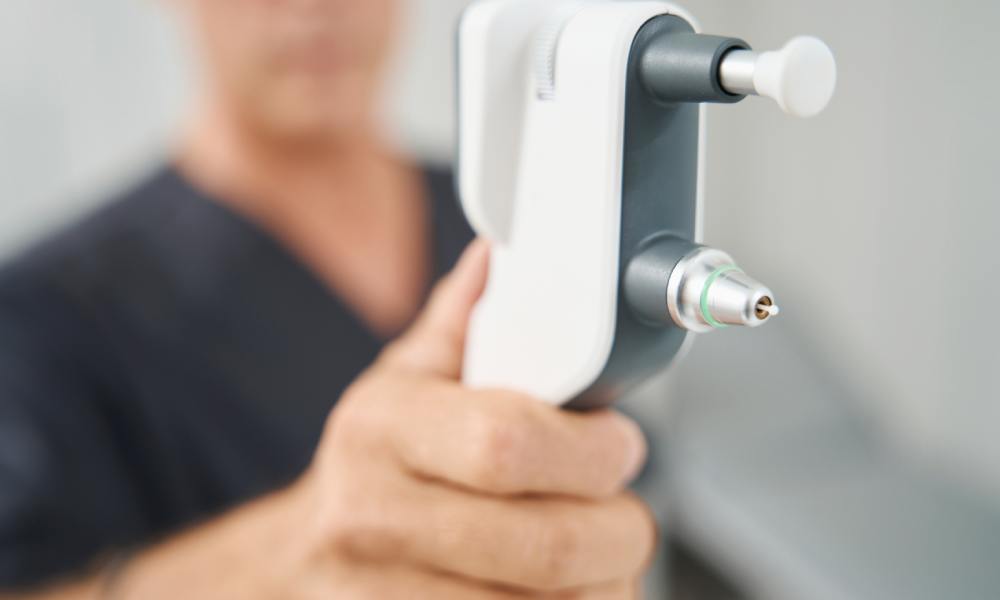 A close-up of a white and gray tonometer device. A white male wearing a dark colored shirt holds the device with one hand.