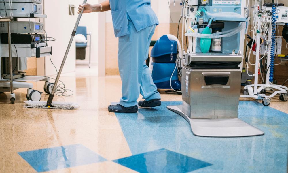 A hospital worker wearing blue scrubs pushes a mop around a hospital room, full of essential lifesaving machines and cords.