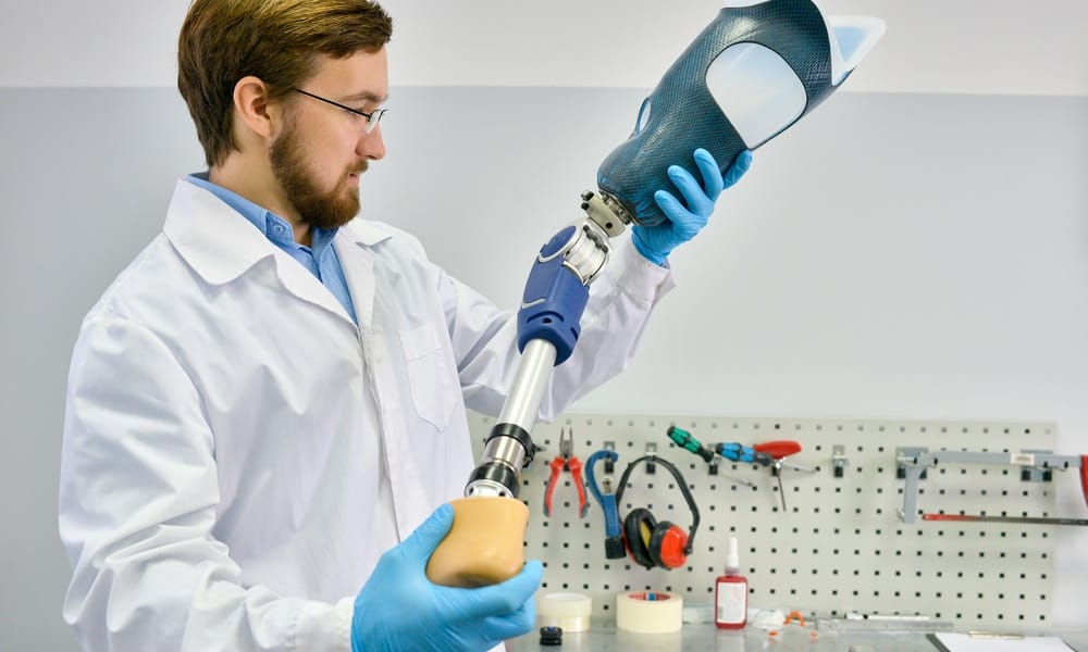 A male prosthetics technician wearing a lab coat holds a leg prosthetic with a flexible knee joint in a lab.