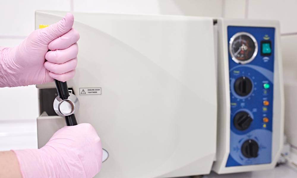 A medical worker wearing pink latex gloves closes the door to an autoclave chamber and uses both hands to seal the lock.