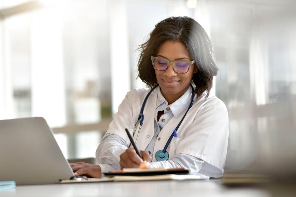 A smiling female healthcare professional is writing on a clipboard with one hand while typing on a laptop with the other.