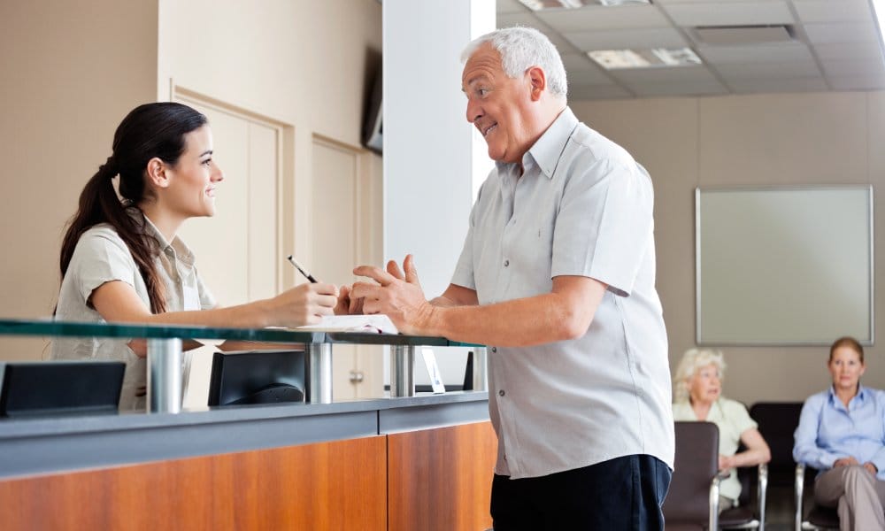 A man communicating with a female receptionist. There are two older women in the background waiting for their turn.