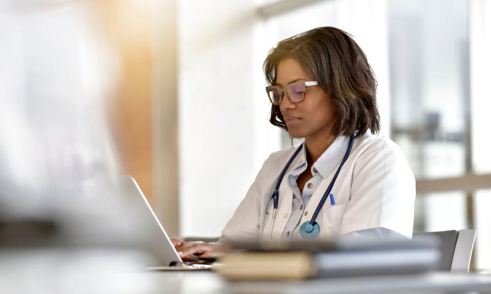 A female healthcare professional sits at a white desk typing on a silver, open laptop. She's wearing a lab coat.