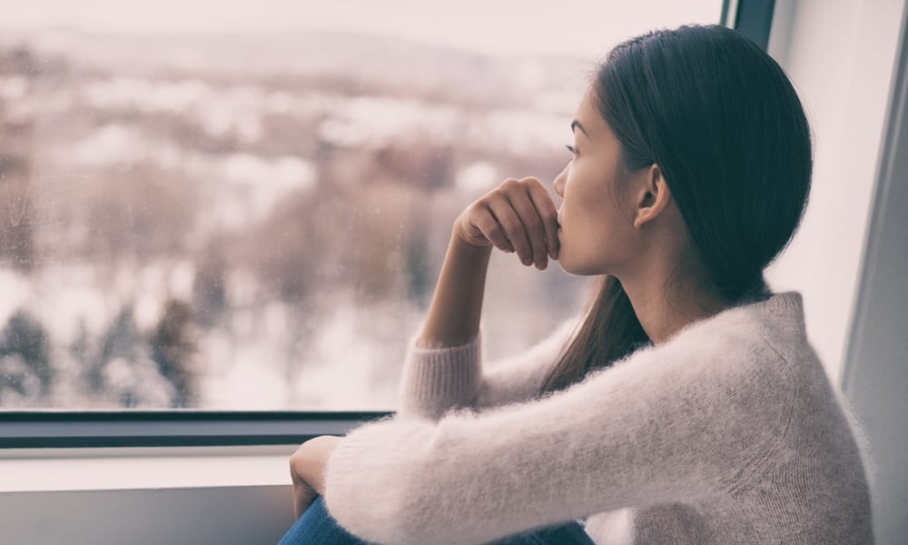 A woman, wearing a fuzzy sweater, is thinking while looking out the window. It's cold and snowing outside.