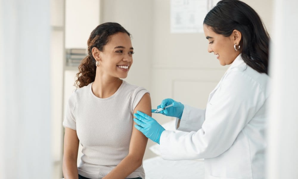 A woman smiles as a doctor gives her an injection in her upper arm. They are in a brightly lit medical exam room.