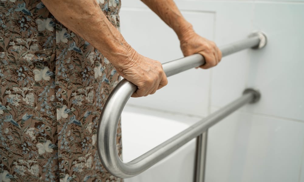 A close-up of an elderly person using a grab bar in a bathroom. Both of their hands are on the bar and they are standing.