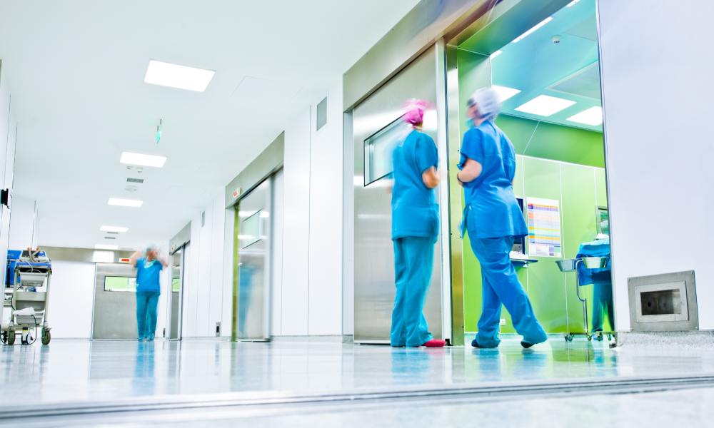 Several doctors in blue scrubs and face masks wander the hallways of a hospital, with fluorescent lights illuminating.