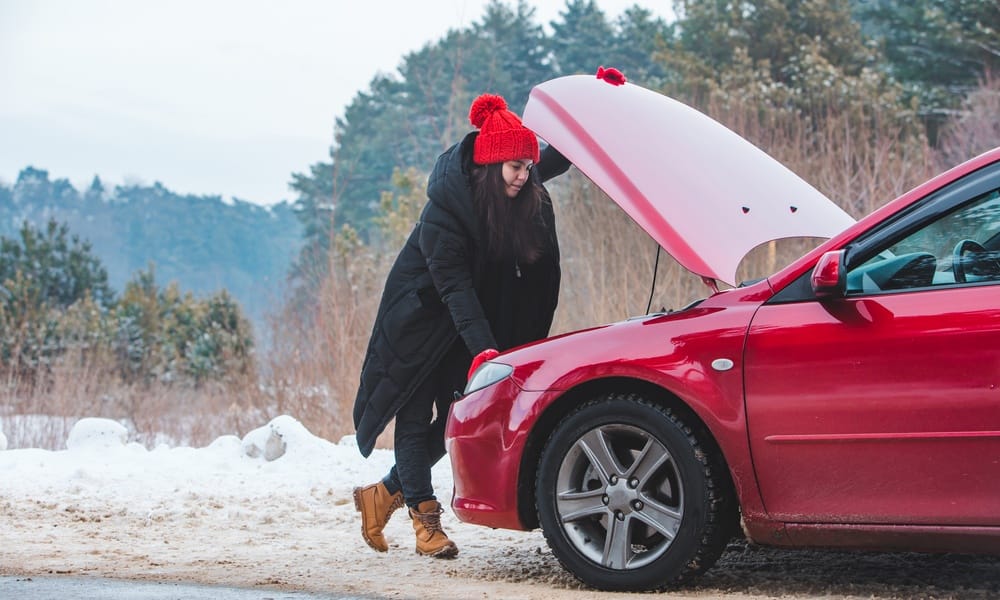 A woman looking under the hood of a car on the side of the road. She has a coat on, and there is snow on the ground.