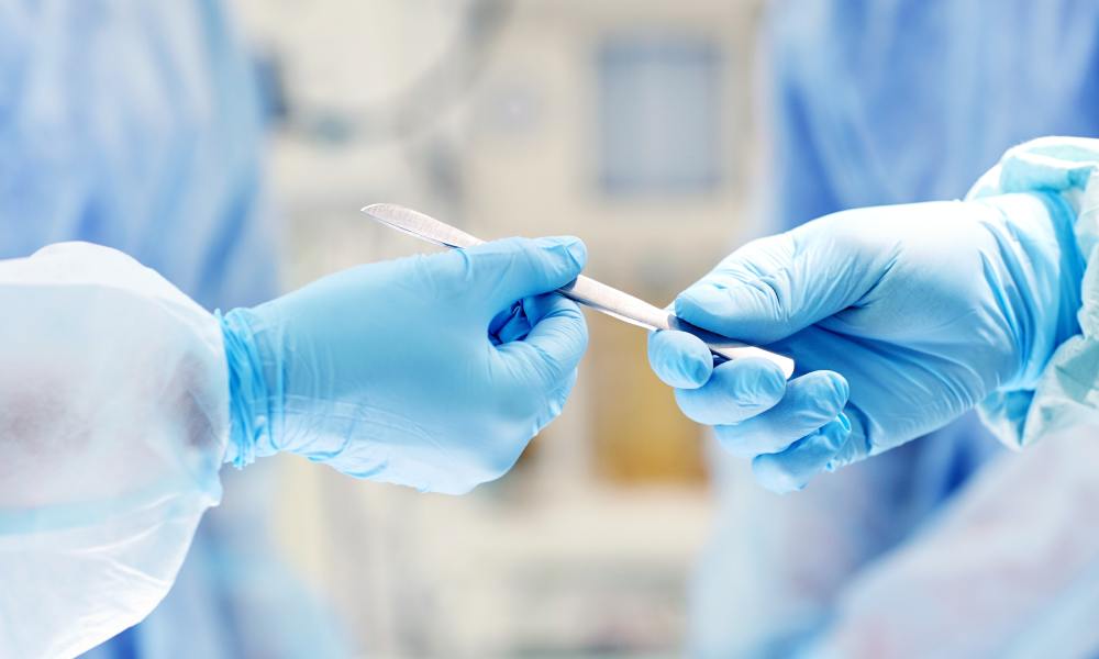 A close-up of gloved hands in gowns exchanging a scalpel in a sterile medical environment, with a blurred background.