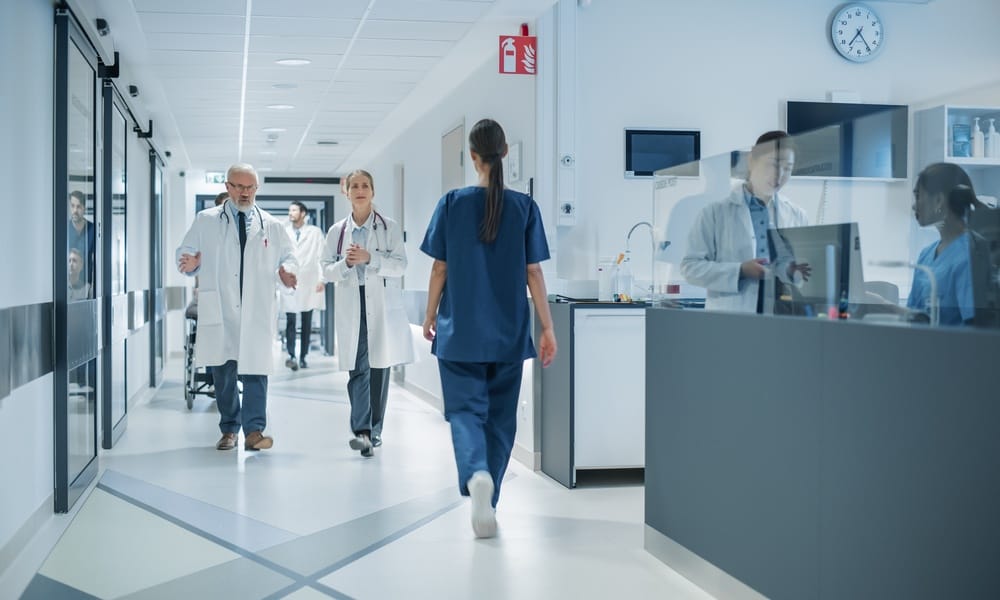 A busy hospital hallway with doctors and nurses walking, a reception desk on the right, and glass doors on the left.