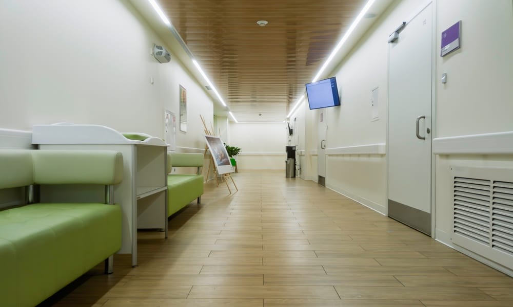 The hallway of a healthcare facility has light bars installed at the cornice on both sides. Green sofas line the hall.