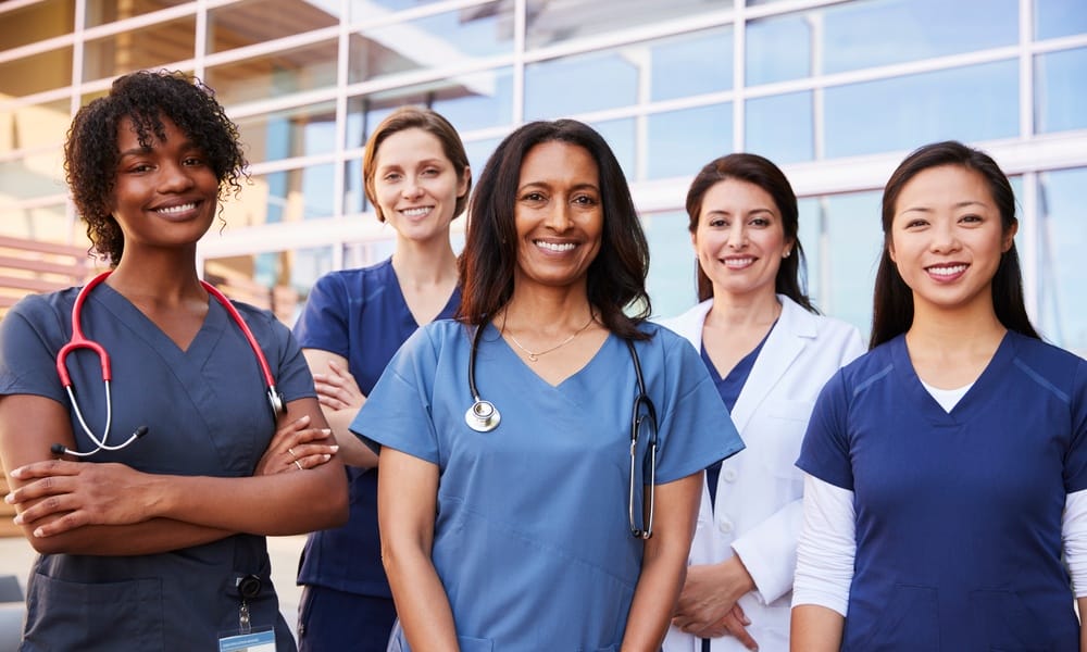 A group of five women, all wearing blue scrubs, standing and smiling in front of a wall of glass inside a building.