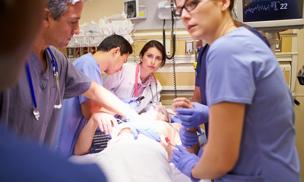 Six healthcare professionals are in a hospital room with a patient. The patient is wearing an oxygen mask.